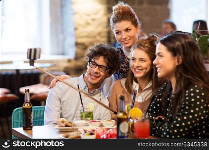 leisure, technology, friendship, people and holidays concept - happy friends with drinks and taking picture by smartphone selfie stick at bar or cafe. friends taking selfie by smartphone at bar or cafe