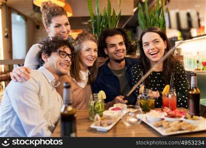 leisure, technology, friendship, people and holidays concept - happy friends with food and drinks taking picture by smartphone selfie stick at bar or cafe. friends taking selfie by smartphone at bar or cafe