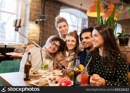 leisure, technology, friendship, people and holidays concept - happy friends with food and drinks taking picture by smartphone selfie stick at bar or cafe. friends taking selfie by smartphone at bar or cafe