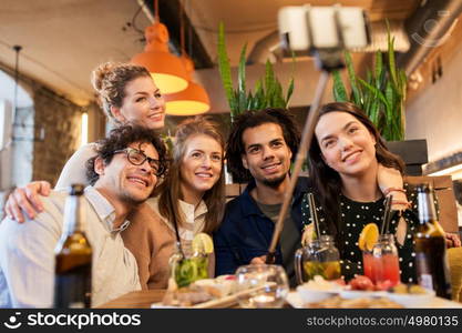 leisure, technology, friendship, people and holidays concept - happy friends with food and drinks taking picture by smartphone selfie stick at bar or cafe. friends taking selfie by smartphone at bar or cafe