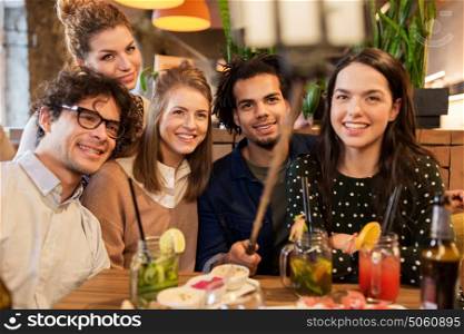 leisure, technology, friendship, people and holidays concept - happy friends with food and drinks and taking picture by smartphone selfie stick at bar or cafe. friends taking selfie by smartphone at bar or cafe