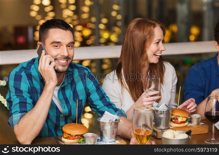 leisure, technology, lifestyle and people concept - happy man with smartphone and friends dining at restaurant