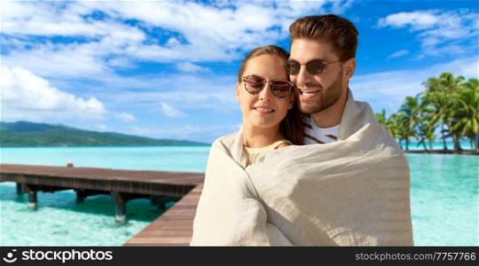 leisure, travel and tourism concept - happy couple in sunglasses covered with blanket hugging over tropical beach background in french polynesia. happy couple covered with blanket hugging on beach