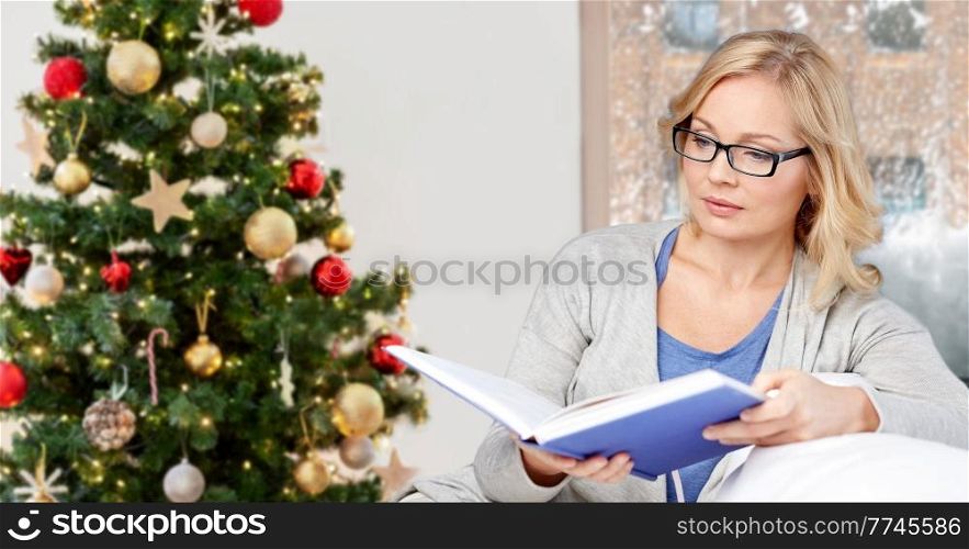 leisure, winter holidays and people concept - middle aged woman reading book and sitting on couch at home over christmas tree lights on background. woman reading book at home on christmas