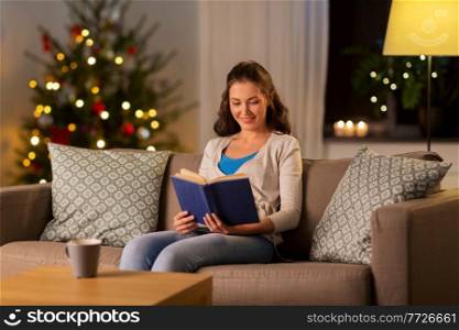 leisure, winter holidays and people concept - young woman reading book at home in evening over christmas tree lights on background. young woman reading book at home on christmas
