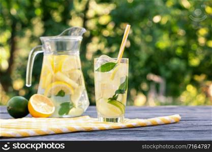 Lemonade in jug and glass and lemon with lime on wooden table. Natural green background. Summer refreshing drink. Lemonade in jug and glass and lemon with lime on wooden table