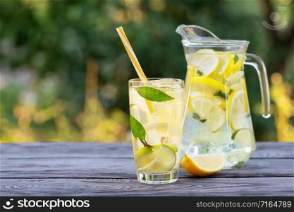 Lemonade in jug and glass and slice of lemon on wooden table. Natural green background. Summer refreshing drink. Lemonade in jug and glass and slice of lemon on wooden table