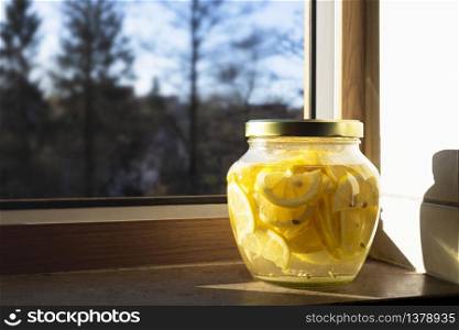 Lemonade with slices of lemons in a big jar on the kitchen window sill in the morning sunlight. Jar on the window sill with homemade lemonade. Healthy drink made with organic lemons, used for body detoxification and diet