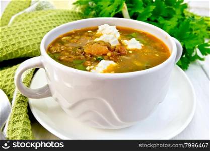 Lentil soup with spinach, tomatoes and feta cheese in a white bowl, spoon on a green napkin, parsley on the background light wooden boards