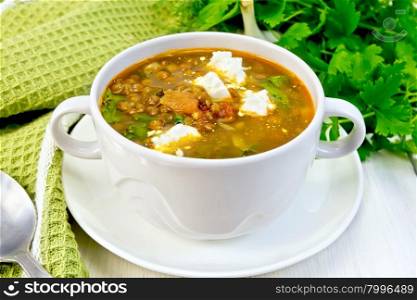 Lentil soup with spinach, tomatoes and feta cheese in a white bowl, spoon on a kitchen towel, parsley on the background light wooden boards