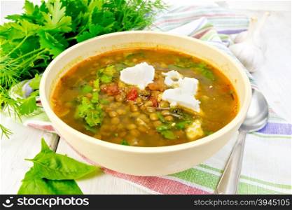 Lentil soup with spinach, tomatoes and feta cheese in a yellow bowl, spoon on a kitchen towel, parsley and spinach on the background light wooden boards