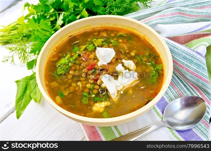 Lentil soup with spinach, tomatoes and feta cheese in a yellow bowl, spoon on a napkin, parsley and spinach on the background light wooden boards