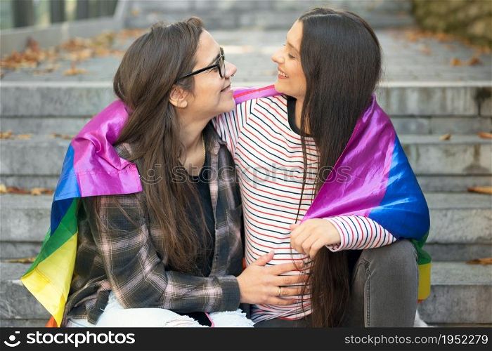 Lesbian couple sitting on steps hugging with rainbow flag at urban scenery. High quality 4k footage. Lesbian couple sitting on steps hugging with rainbow flag at urban scenery.