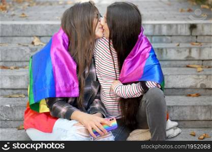 Lesbian couple sitting on steps with rainbow flag, hugging and kissing at urban scenery. High quality photo.. Lesbian couple sitting on steps with rainbow flag, hugging and kissing at urban scenery.