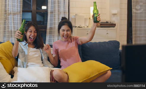 Lesbian lgbt women couple party at home, Asian female drinking beer watching TV cheer soccer funny moment together on sofa in living room in night. Young lover football fan, celebrate holiday concept.
