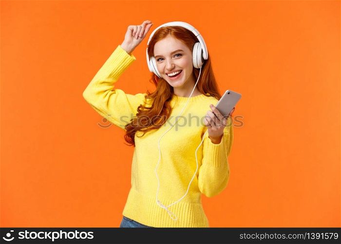 Lets dance. Cheerful carefree modern hipster girl with red curly hair and freckles, dancing lift hand up and holding smartphone, listen music with new ordered online headphones, orange background.. Lets dance. Cheerful carefree modern hipster girl with red curly hair and freckles, dancing lift hand up and holding smartphone, listen music with new ordered online headphones, orange background