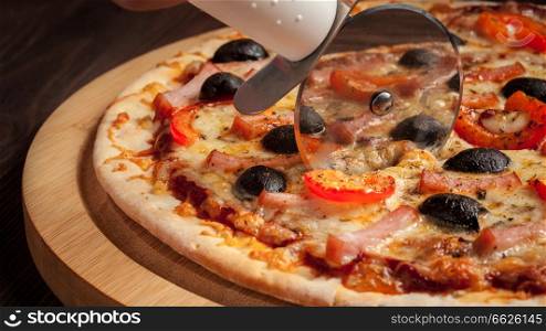 Letterbox panorama of pizza cutter (wheel) slicing ham pizza with capsicum and olives on wooden board on table. Pizza cutter wheel slicing ham pizza