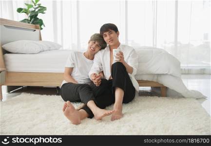 LGBT Asian attractive gay couple sitting on the floor and holding hands spend time together in the morning at bedroom and looking at camera