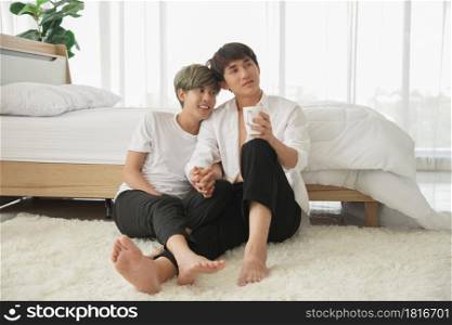LGBT Asian attractive gay couple sitting on the floor and holding hands spend time together in the morning at bedroom and looking outside
