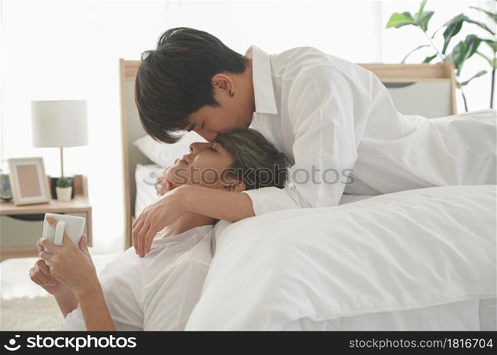 LGBT Asian handsome young gay couple expression love, kissing forehead lying on bed and holding cup of coffee in the morning