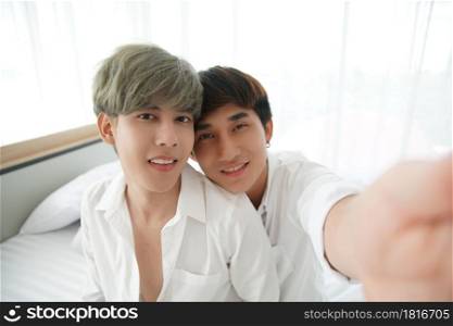 LGBT Asian young gay couple taking a selfie photo smiling and looking at camera together at bedroom