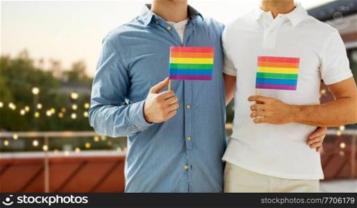 lgbt, homosexuality and people concept - close up of happy male gay couple hugging and holding rainbow flags over roof top party background. male gay couple hugging and holding rainbow flags