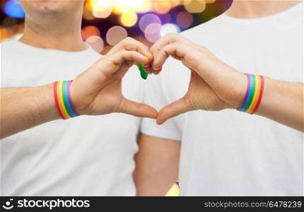 lgbt, same-sex love and homosexual relationships concept - close up of happy male couple wearing gay pride rainbow awareness wristbands showing hand heart gesture over lights background. gay couple with rainbow wristbands and hand heart. gay couple with rainbow wristbands and hand heart