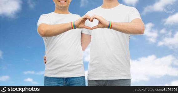lgbt, same-sex love and homosexual relationships concept - close up of happy smiling hugging male couple wearing gay pride rainbow awareness wristbands showing hand heart gesture. couple with gay pride rainbow wristbands and heart. couple with gay pride rainbow wristbands and heart