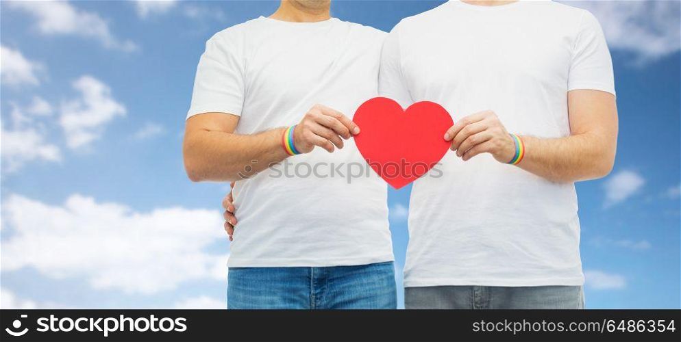 lgbt, same-sex love and homosexual relationships concept - close up of hugging male couple wearing gay pride rainbow awareness wristbands holding red heart shape over blue sky and clouds background. couple with gay pride rainbow wristbands and heart. couple with gay pride rainbow wristbands and heart