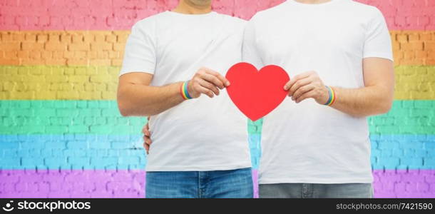 lgbt, same-sex love and homosexual relationships concept - close up of hugging male couple wearing gay pride awareness wristbands holding red heart shape over rainbow colored brick wall background. couple with gay pride rainbow wristbands and heart