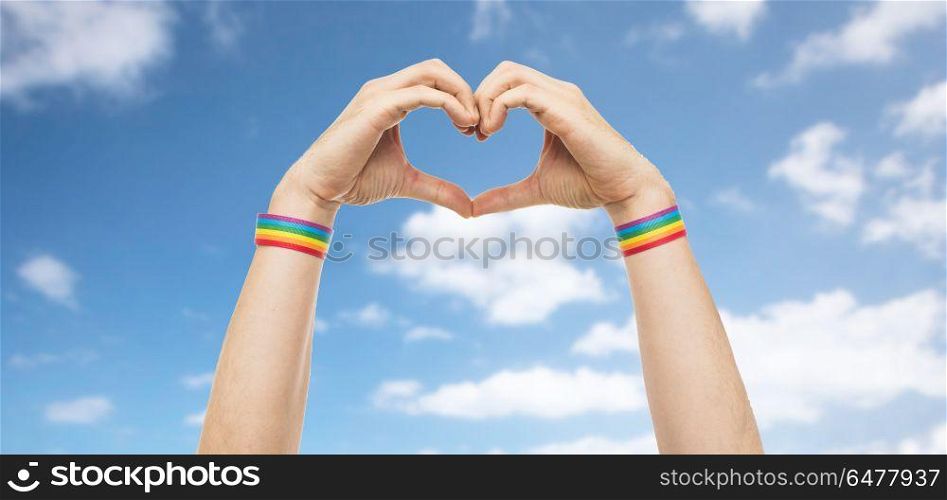 lgbt, same-sex love and homosexual relationships concept - close up of male hands with gay pride rainbow awareness wristbands showing heart gesture over blue sky and clouds background. male hands with gay pride wristbands showing heart. male hands with gay pride wristbands showing heart