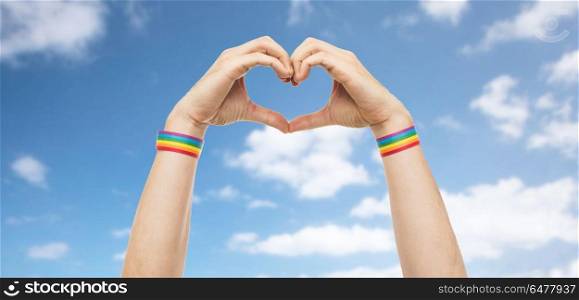 lgbt, same-sex love and homosexual relationships concept - close up of male hands with gay pride rainbow awareness wristbands showing heart gesture over blue sky and clouds background. male hands with gay pride wristbands showing heart. male hands with gay pride wristbands showing heart