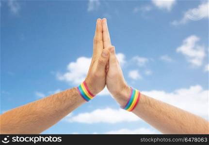 lgbt, same-sex love and homosexual relationships concept - close up of male couple hands with gay pride rainbow awareness wristbands making high five gesture over blue sky and clouds background. hands with gay pride wristbands make high five. hands with gay pride wristbands make high five