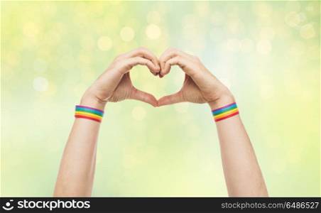 lgbt, same-sex love and homosexual relationships concept - close up of male hands with gay pride rainbow awareness wristbands showing heart gesture over green lights background. male hands with gay pride wristbands showing heart. male hands with gay pride wristbands showing heart