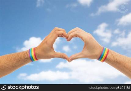 lgbt, same-sex love and homosexual relationships concept - close up of male couple hands with gay pride rainbow awareness wristbands showing heart gesture over blue sky and clouds background. male hands with gay pride wristbands showing heart. male hands with gay pride wristbands showing heart