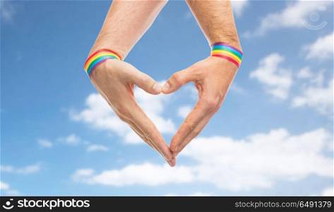 lgbt, same-sex love and homosexual relationships concept - close up of male couple hands with gay pride rainbow awareness wristbands showing heart gesture over blue sky and clouds background. male hands with gay pride wristbands showing heart. male hands with gay pride wristbands showing heart