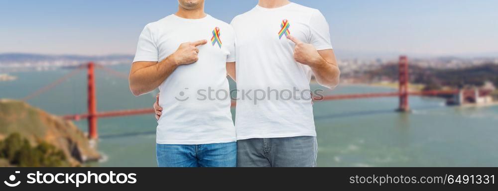 lgbt, same-sex relationships and homosexual concept - close up of happy male couple pointing at gay pride rainbow awareness ribbons on chest over golden gate bridge in san francisco bay background. close up of couple with gay pride rainbow ribbons. close up of couple with gay pride rainbow ribbons
