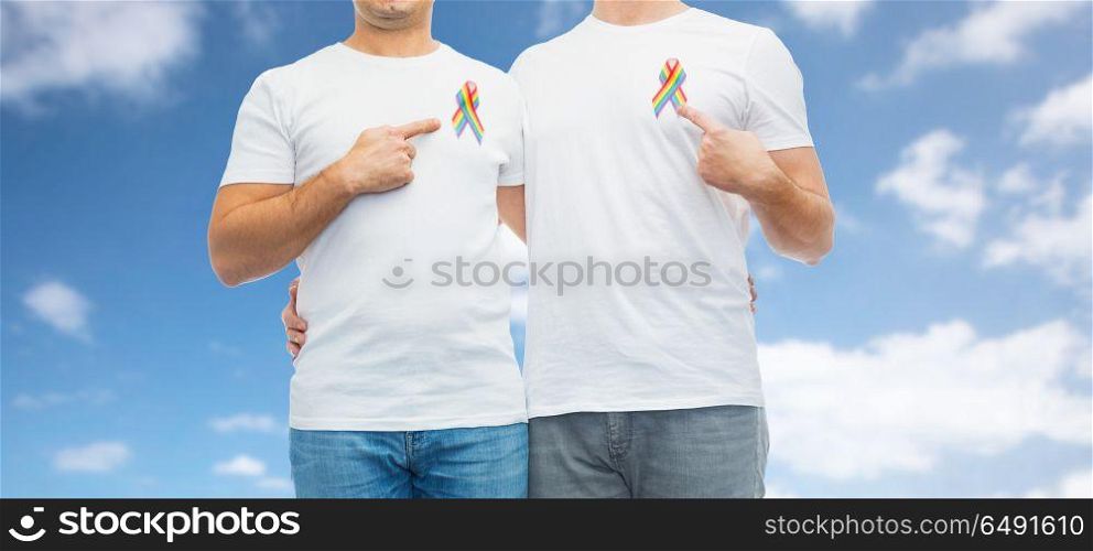 lgbt, same-sex relationships and homosexual concept - close up of happy male couple pointing at gay pride rainbow awareness ribbons on chest over blue sky and clouds background. close up of couple with gay pride rainbow ribbons. close up of couple with gay pride rainbow ribbons