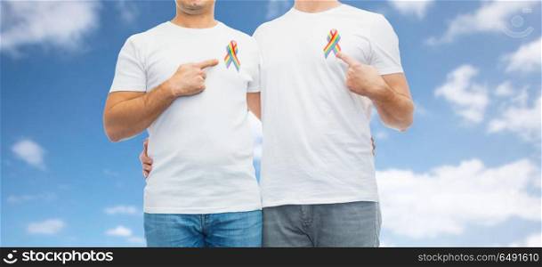 lgbt, same-sex relationships and homosexual concept - close up of happy male couple pointing at gay pride rainbow awareness ribbons on chest over blue sky and clouds background. close up of couple with gay pride rainbow ribbons. close up of couple with gay pride rainbow ribbons