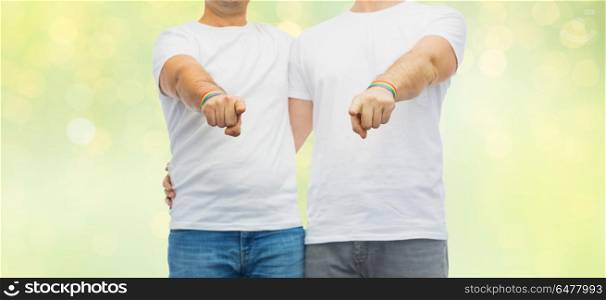 lgbt, same-sex relationships and homosexual concept - close up of hugging male couple wearing gay pride rainbow awareness wristbands and pointing fingers at you over green lights background. couple with gay pride rainbow wristbands. couple with gay pride rainbow wristbands