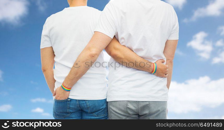 lgbt, same-sex relationships and homosexual concept - close up of hugging male couple wearing rainbow gay pride awareness wristbands over blue sky and clouds background. male couple with gay pride rainbow wristbands. male couple with gay pride rainbow wristbands