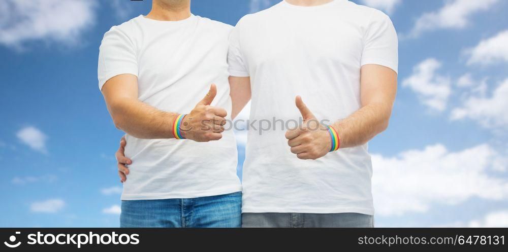 lgbt, same-sex relationships and homosexual concept - close up of male couple wearing gay pride rainbow awareness wristbands and showing thumbs up over blue sky and clouds background. gay couple with rainbow wristbands shows thumbs up. gay couple with rainbow wristbands shows thumbs up