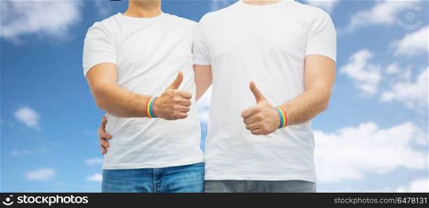 lgbt, same-sex relationships and homosexual concept - close up of male couple wearing gay pride rainbow awareness wristbands and showing thumbs up over blue sky and clouds background. gay couple with rainbow wristbands shows thumbs up. gay couple with rainbow wristbands shows thumbs up