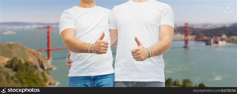 lgbt, same-sex relationships and homosexual concept - close up of male couple with gay pride rainbow awareness wristbands and showing thumbs up over golden gate bridge in san francisco bay background. gay couple with rainbow wristbands shows thumbs up. gay couple with rainbow wristbands shows thumbs up