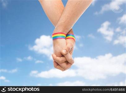 lgbt, same-sex relationships and homosexual concept - close up of male couple wearing gay pride rainbow awareness wristbands holding hands over blue sky and clouds background. hands of couple with gay pride rainbow wristbands. hands of couple with gay pride rainbow wristbands