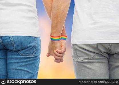 lgbt, same-sex relationships and homosexual concept - close up of male couple wearing gay pride rainbow awareness wristbands holding hands over evening sky background. male couple with gay pride rainbow wristbands