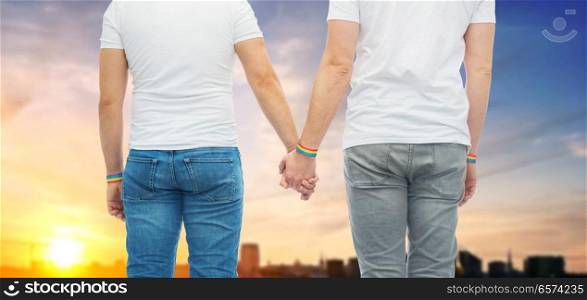 lgbt, same-sex relationships and homosexual concept - close up of male couple wearing gay pride rainbow awareness wristbands holding hands over sunset in tallinn city background. male couple with gay pride rainbow wristbands