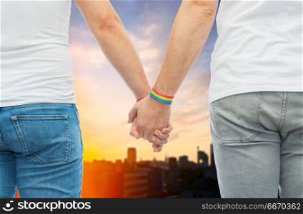 lgbt, same-sex relationships and homosexual concept - close up of male couple wearing gay pride rainbow awareness wristbands holding hands over city sunset background. male couple with gay pride rainbow wristbands. male couple with gay pride rainbow wristbands