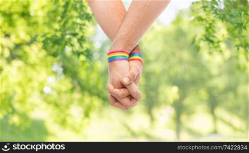 lgbt, same-sex relationships and homosexual concept - close up of male couple wearing gay pride rainbow awareness wristbands holding hands over green natural background. hands of couple with gay pride rainbow wristbands