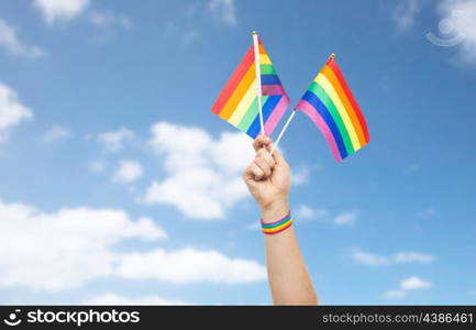 lgbt, same-sex relationships and homosexual concept - close up of male hand wearing gay pride awareness wristband holding rainbow flags over blue sky and clouds background. hand with gay pride rainbow flags and wristband. hand with gay pride rainbow flags and wristband